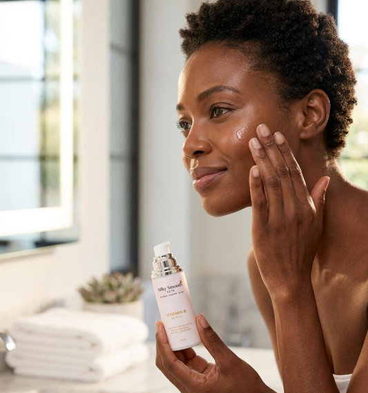 Woman applying Vitamin B serum to her face in a bright bathroom, holding a bottle labeled "Silky Smooth