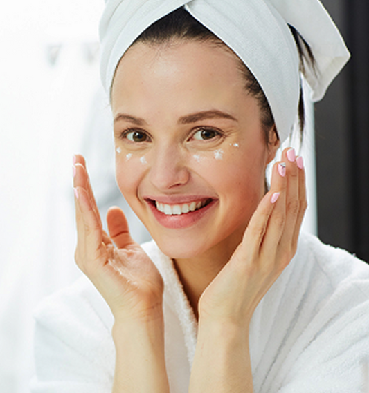 Smiling woman in a white robe and towel applies eye cream, enhancing her skincare routine with a fresh, radiant glow.